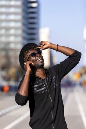 Laughing Black man wearing hat & sunglasses standing on the street talking on cellphone