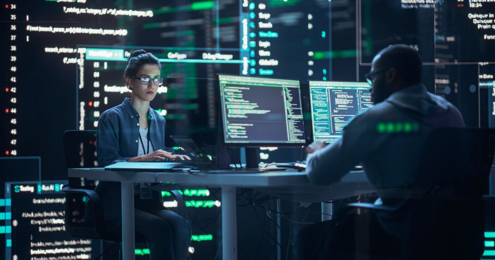Diverse Young Man and Woman Working on Desktop Computers, Surrounded by Big Screens Displaying Code Lines.