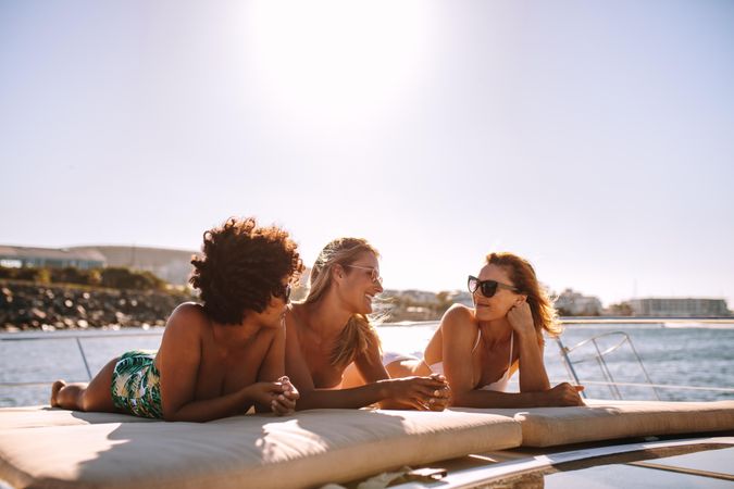 Smiling female friends wearing swimsuits relaxing on yacht deck and chatting on summer day