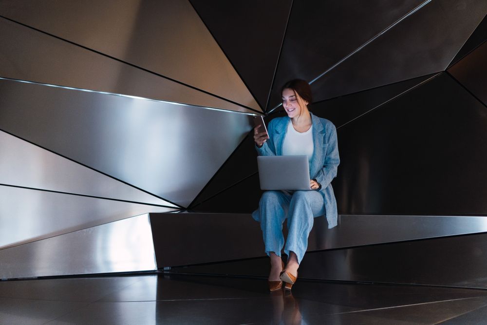 Cheerful woman with laptop browsing smartphone in futuristic studio