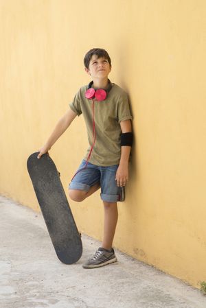 Young boy leaning on a yellow wall with headphones on neck, holding a skateboard while looking up