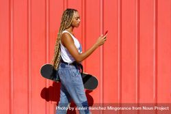 Woman walking in front of red wall texting on phone and holding a skateboard 5XygQ4