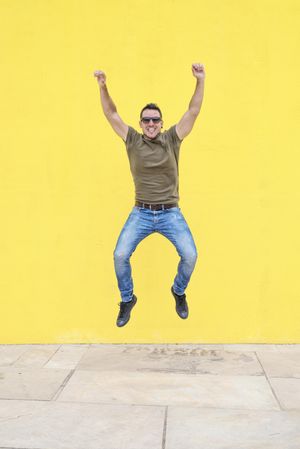 Happy male jumping outside in front of yellow wall with arms up