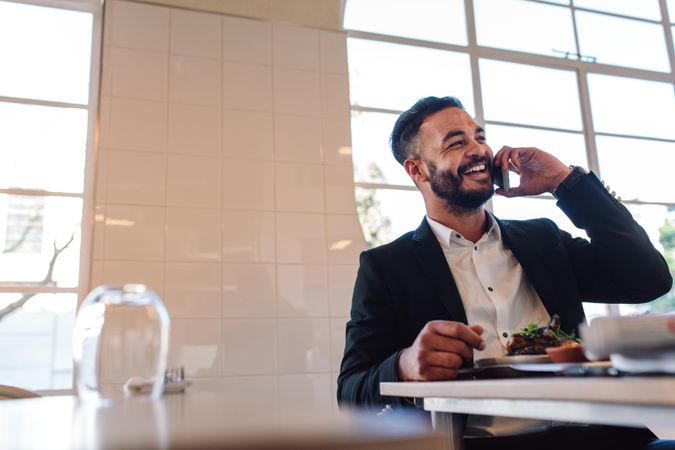 Smiling young business man sitting at restaurant table with food and talking on mobile phone