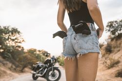 Woman putting on driving gloves while standing on road beside motorbike