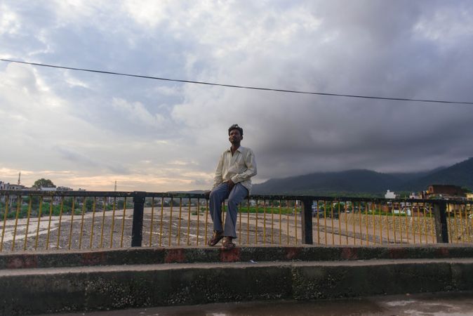 Indian man sitting on railing of bridge over shallow river on cloudy day in Dehradun, India