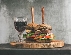 Two cheeseburgers skewered with knives, stacked with fresh vegetables, on wooden board, with wine
