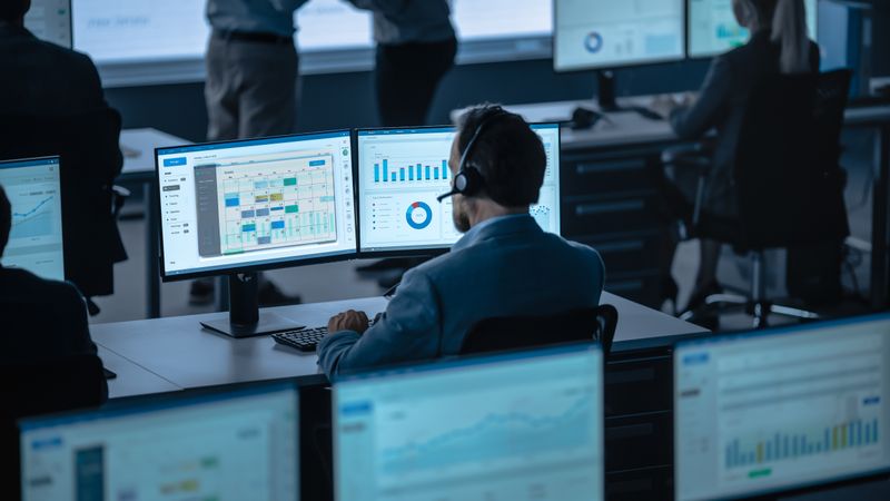 Analyst Reviews Scheduling and Performance Data on Dual Monitors in Busy Office.