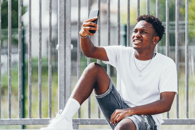Sportive black men thoughtfully browsing smartphone while sitting on fence