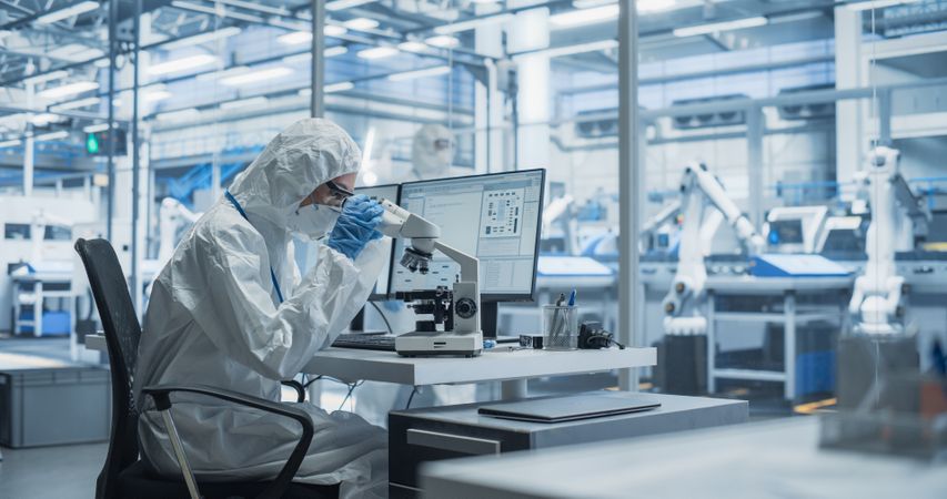 Modern Factory: Industrial Engineer Working on Desktop Computer in Clean Sterile Coveralls. Scientist Using a Microscope, Developing Advanced Solutions for High-Tech Medical Vaccine and Gene Research