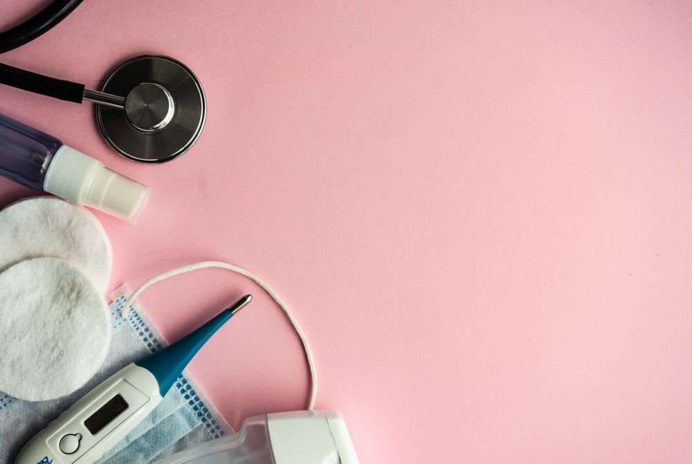 Top view of pink table with facemask, cotton, spray, and digital thermometer
