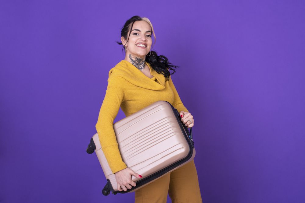 happy woman traveler holding her suitcase against purple background.