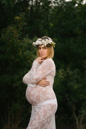 Pregnant woman with closed eyes in white lace dress touching chin while standing in forest