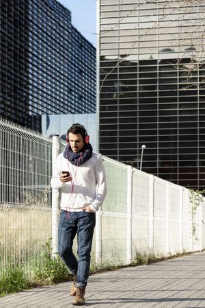 Young man walking in scarf past metallic fence and holding smartphone outdoors