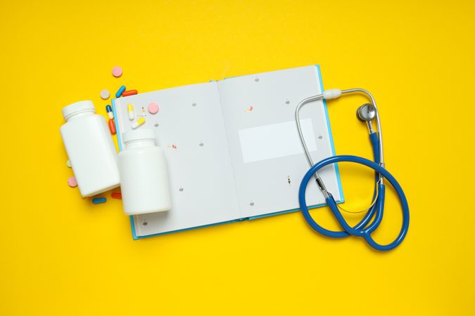 Notepad, stethoscope and jars of pills on yellow background, top view