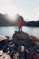 Back view of man in red jacket standing on rock by lake waterside