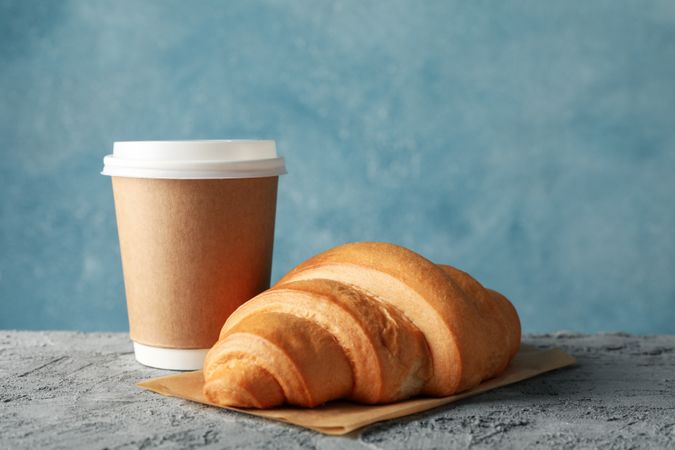 Paper cup of coffee and croissant on grey table
