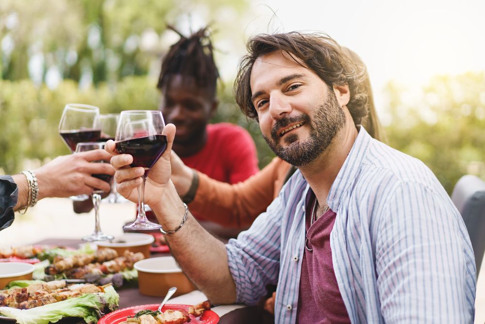 Man looking at camera celebrating with multi-ethnic friends toasting with red wine at picnic table