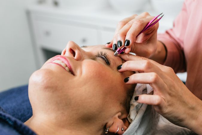 Unrecognizable beautician plucking eyebrows on face of woman in salon