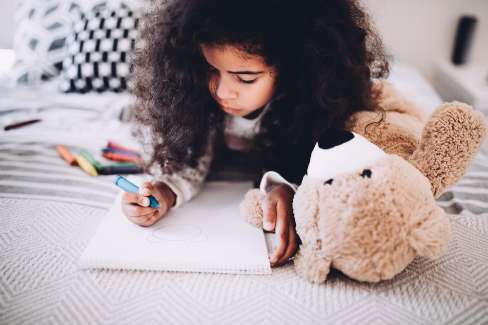Little girl laying on her bed drawing in her sketchbook with crayons