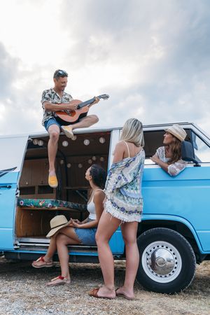 Man playing on guitar on car roof and women listening with pleasure