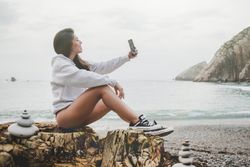 Woman taking selfie on smartphone on shore near stones and water