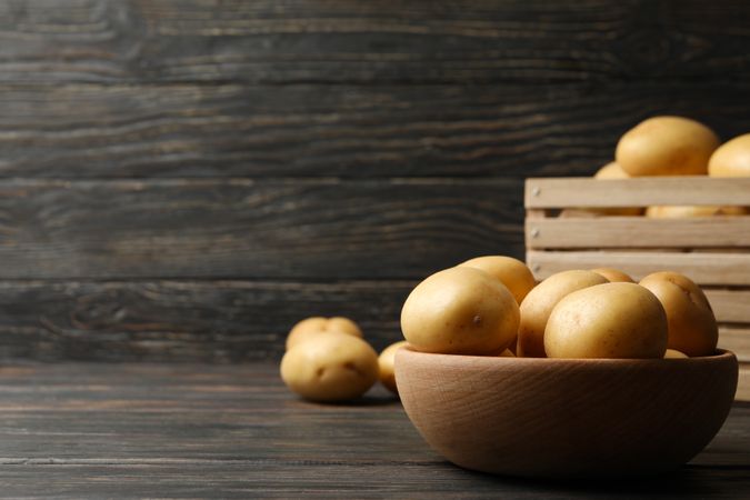 Side view of bowl of potatoes next to basket of potatoes on dark table, copy space