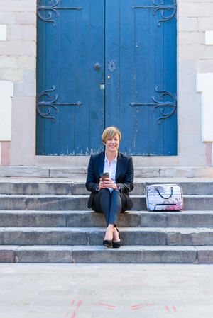 Smiling female in blazer sitting in front of blue door outside with coffee