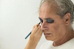 Hand applying dark and blue eyeshadow to middle aged man's eye in studio bGXrv0
