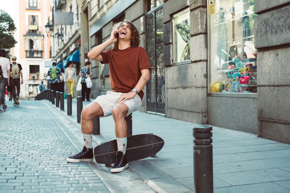 Amused man with longboard talking on phone while resting on street