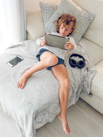 Concentrated preteen boy lying on sofa and using tablet