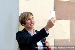 Smiling woman wearing a blazer and taking photo with phone outside bGprl0