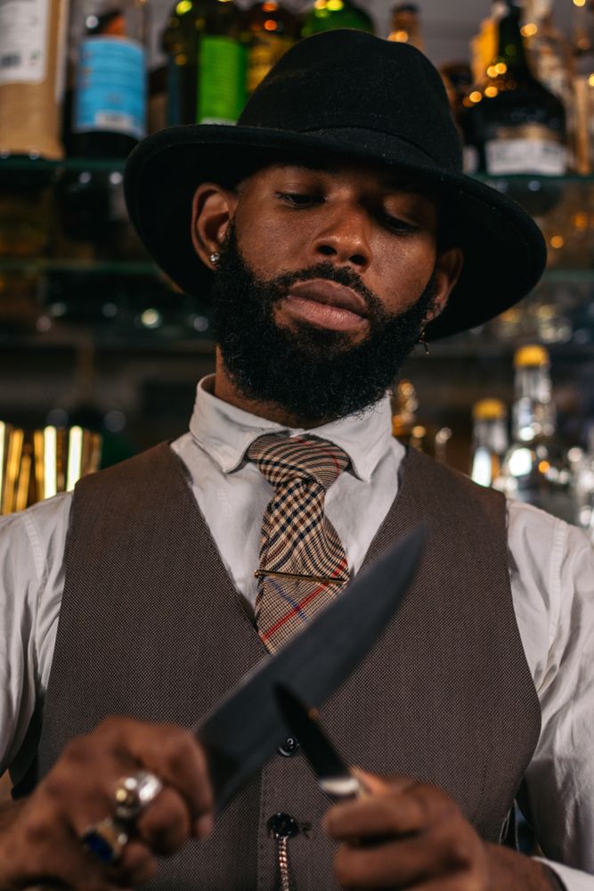 Close-up of a stylish Black bartender in a hat sharpening his knives