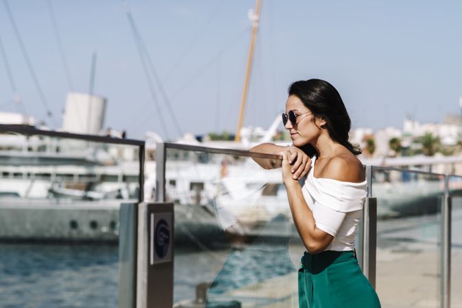 Side view of dreamy young woman with sunglasses leaning on fence and looking away on harbor dock