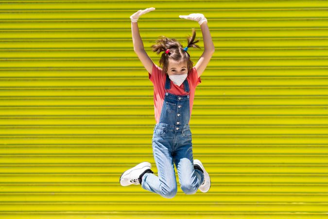 Cute girl jumping in protective mask against a yellow wall