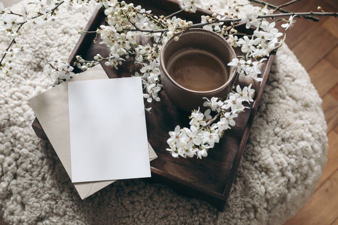 Spring still life composition. Greeting card mockup, cup of coffee. Feminine styled photo. Floral scene with blurred cherry tree blossoms on wool taburet, stool. Wooden parquet floor. Selective focus