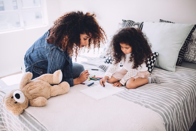 Little girl and her mother sitting on a bed drawing in a sketch book using crayons