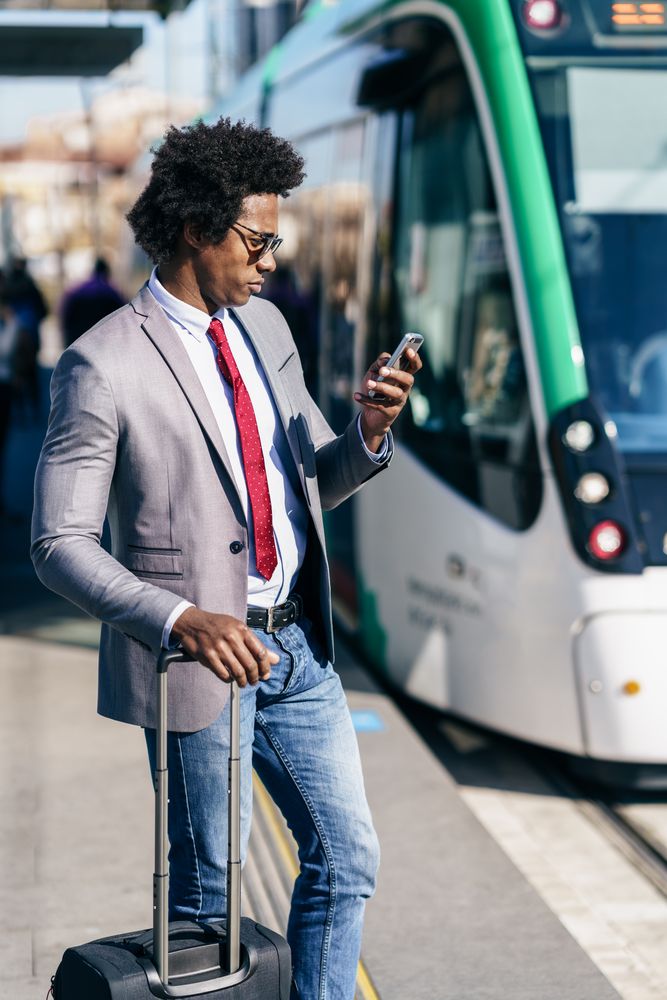 Man in jeans and dress shirt with tie standing on train platform with roller suitcase