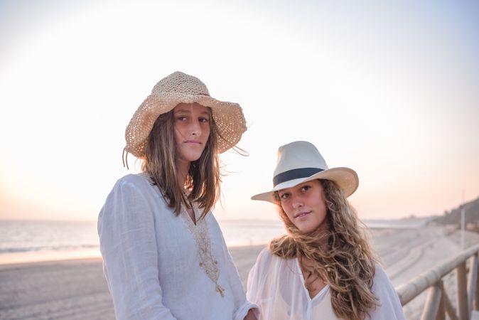 Smiling girlfriends in hats talking sitting on wooden fence on beach