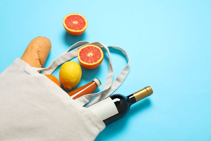 Eco bag with bottle of wine, juice, fruits and baguette on color background, space for text