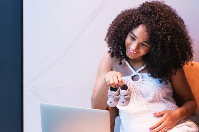 Smiling pregnant Black woman showing baby booties to laptop
