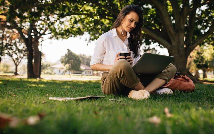 Beautiful woman sitting on grass lawn at park holding a coffee and looking at laptop computer