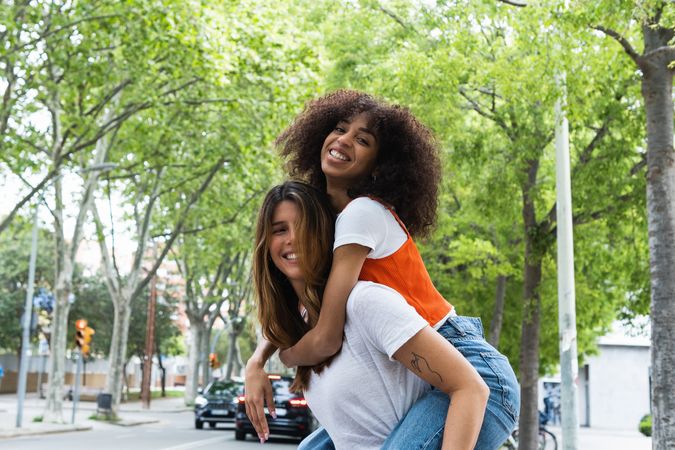 Black woman riding piggyback on smiling girlfriend on road