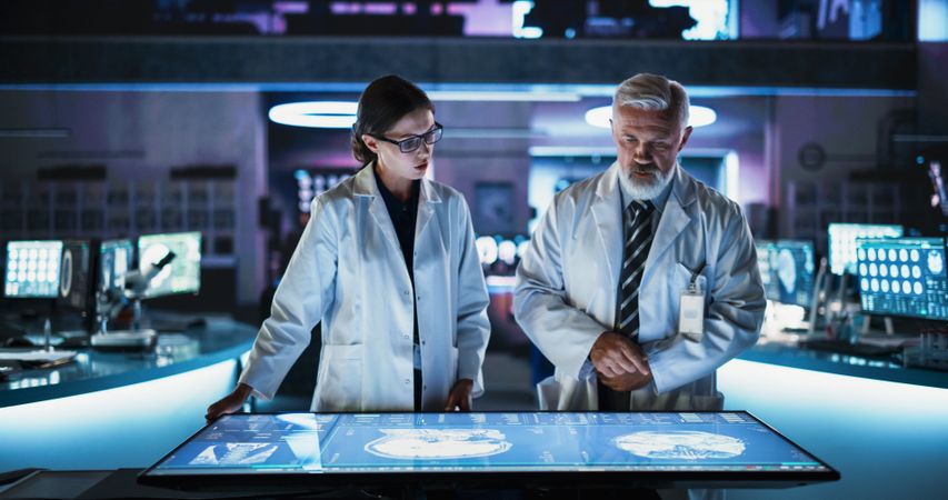 Female Neuroscientist And Male Surgeon Talking, Using Interactive Touch Screen Table With CT Scan Of Brain Damage Patient, Developing Solutions.