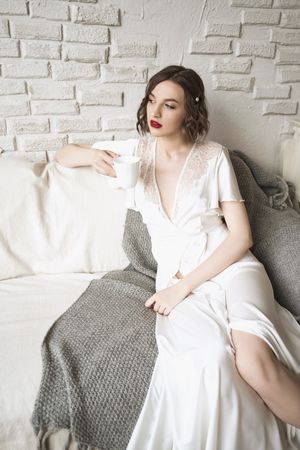 Pensive adult woman looking away and dreaming while sitting on sofa and having hot drink in light modern living room