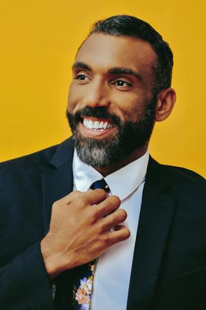 Smiling Black male in suit fixing his floral tie in yellow studio