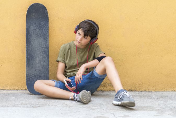 Teenager sitting on ground leaning on a yellow wall while holding a mobile phone