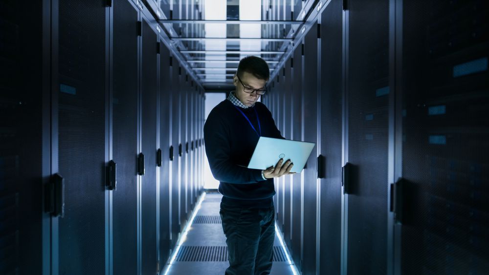 Male IT Engineer Works on a Laptop in front of Server Cabinet at a Big Data Center. Rows of Rack Servers are Seen.