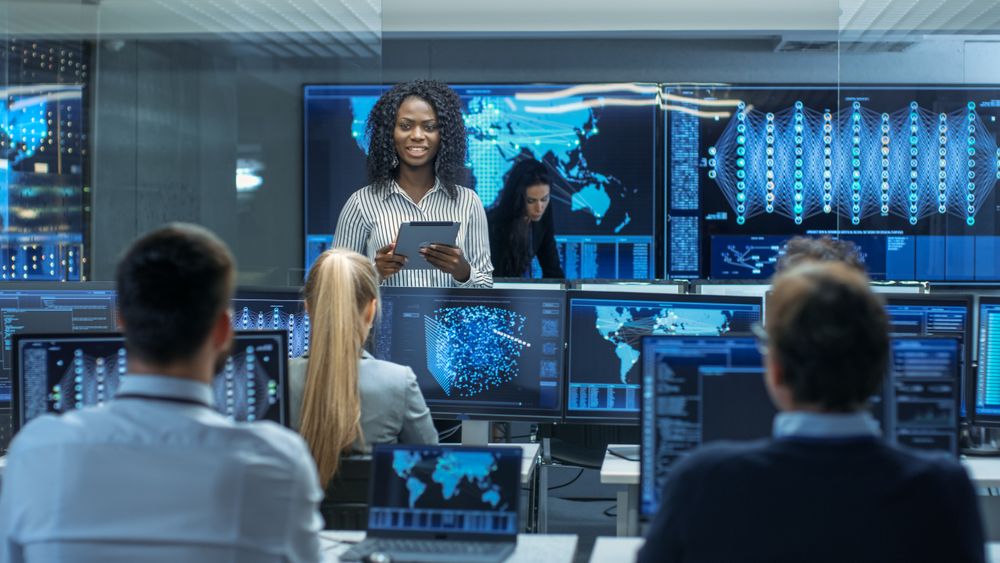 Chief Project Engineer Consults Tablet Computer and Holds Briefing for a Team of Scientists that are Building Machine Learning System.