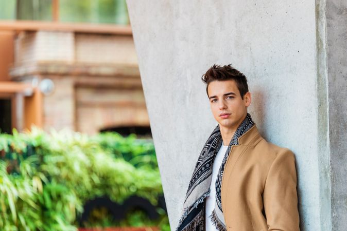 Serious man wearing camel coat and scarf looking at camera against cement wall